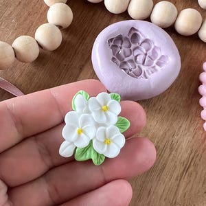 May include: A silicone mold for making small, white flower decorations. The mold is purple and has a detailed design of five flowers with green leaves.