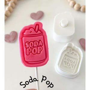 May include: A pink "Soda Pop" shaped lollipop on a stick, with the words "Soda Pop" embossed on the candy. A white plastic mold and a heart-shaped candy are also visible. The word "Soda Pop" is written in cursive.