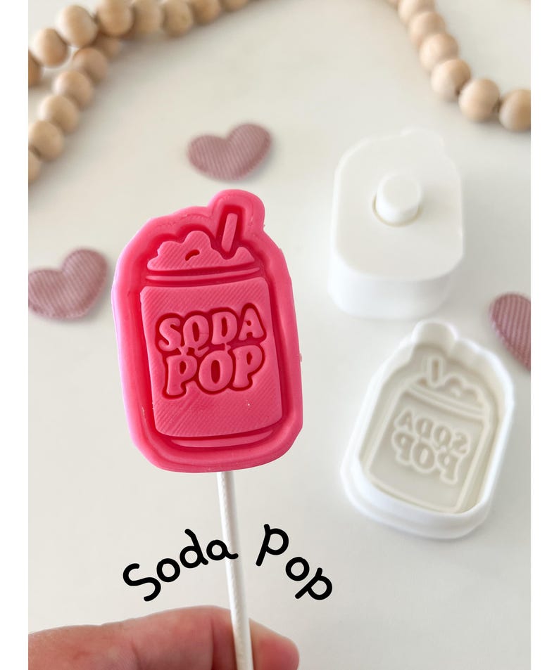 May include: A pink "Soda Pop" shaped lollipop on a stick, with the words "Soda Pop" embossed on the candy. A white plastic mold and a heart-shaped candy are also visible. The word "Soda Pop" is written in cursive.