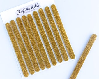 May include: Ten gold glitter craft sticks, each approximately 10 cm long, arranged in a row on a white background. One additional stick is placed horizontally below the row.
