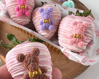 May include: Four chocolate-covered strawberries decorated with pink, purple, and brown rose-shaped teddy bears. Each bear has a gold bow tie.