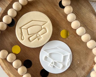 May include: Two white plastic cookie cutters shaped like a graduation cap and diploma. The cutters are on a wooden surface with a string of wooden beads.