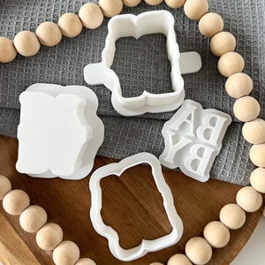 K&ouml;nnte beinhalten: Ein Satz mit vier wei&szlig;en Plastik-Ausstechern in Form der Buchstaben "B", "A", "Y" und einem Rechteck. Die Ausstecher befinden sich auf einer Holzoberfl&auml;che mit einer Schnur aus Holzperlen am Rand.