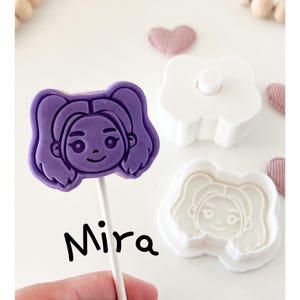 May include: A purple cookie on a stick with a girl's face, labeled "Mira." Next to it are a white cookie cutter and a matching stamp. The background includes wooden beads and pink heart-shaped decorations.