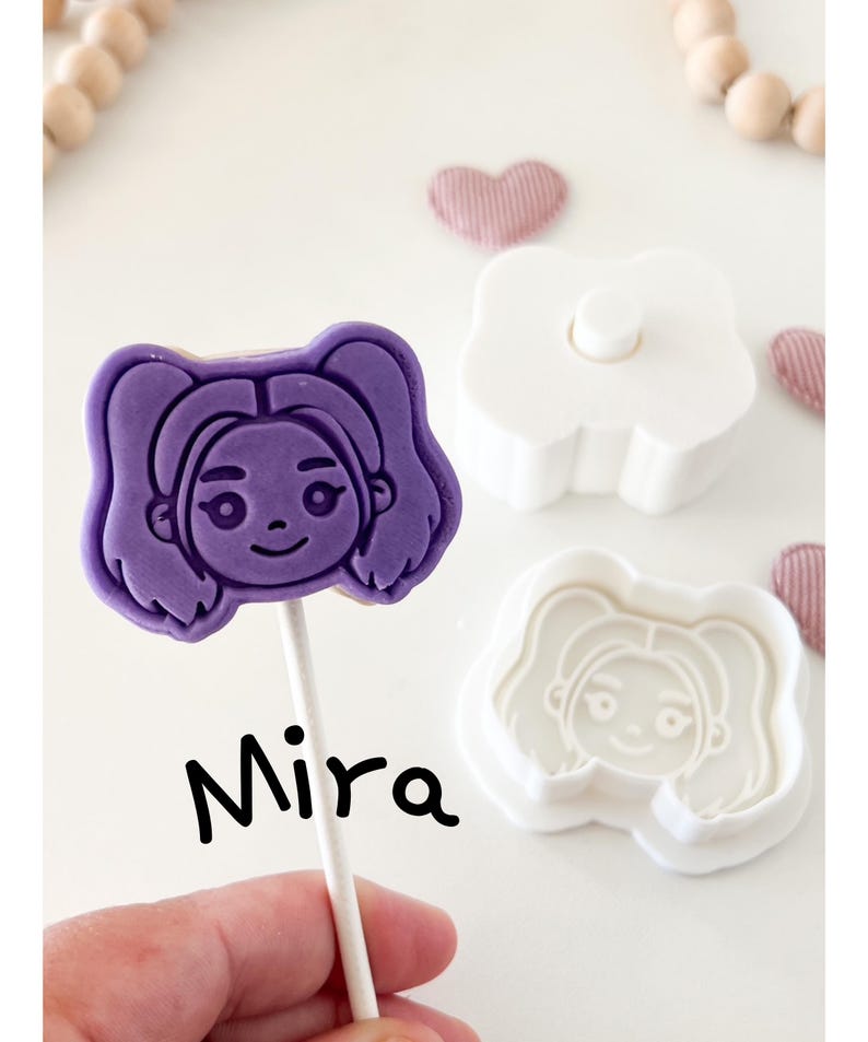 May include: A purple cookie on a stick with a girl's face, labeled "Mira." Next to it are a white cookie cutter and a matching stamp. The background includes wooden beads and pink heart-shaped decorations.