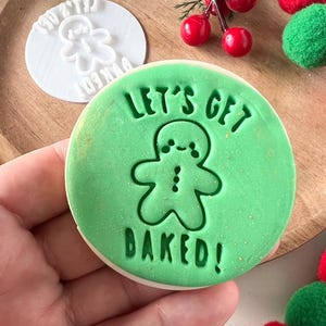 Puede incluir: Una galleta verde con el texto "LET'S GET BAKED!" y un diseño de hombre de jengibre. Es visible un sello de galleta blanco con un diseño de hombre de jengibre. La decoración festiva incluye bayas rojas y pompones verdes.