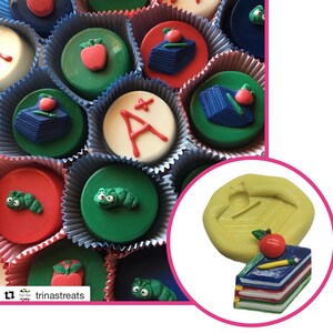 May include: A silicone mold for making chocolate treats shaped like a stack of books with an apple and pencil on top. The mold is yellow and the finished chocolate treats are red, blue, green, and white.