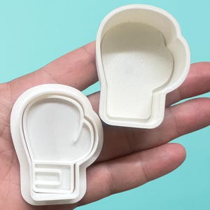 May include: Two white plastic cookie cutters shaped like boxing gloves. The cutters are designed to create cookies with a raised outline of a boxing glove.