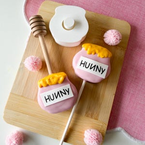 May include: A wooden cutting board holds two pink cake pops shaped like honey pots, topped with yellow frosting and the word "HUUNY". A wooden honey dipper and a white honey pot are also present, along with pink decorative accents.