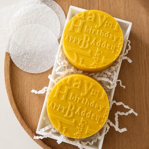 May include: Two circular yellow wax seals featuring embossed text that reads Happy Birthday in a repeating pattern. The seals are nestled in a white rectangular tray filled with white paper shred, accompanied by translucent circular wax paper sheets.