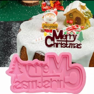Op de afbeelding: Roze siliconen mal voor het bakken van een cake met de woorden "Merry Christmas" in een feestelijk lettertype. De mal wordt getoond op een cake die is versierd met een kerstmanfiguur, een peperkoekhuis en een rode strik.