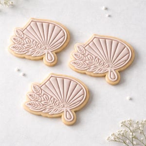May include: Three fan-shaped cookies with a light pink design and a beige border. Each cookie features a detailed fan design with radiating lines and a floral element. The cookies are arranged on a white surface with small white sprinkles.