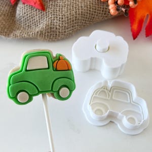 May include: A green truck-shaped cookie on a white stick, topped with an orange pumpkin. Beside it are two white plastic truck-shaped cookie cutters. Autumn-themed treats.