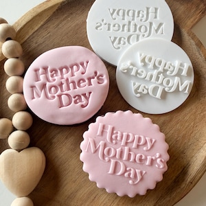 Puede incluir: Una bandeja de madera contiene tres sellos para galletas y dos galletas rosas. Las galletas y los sellos son redondos, con las palabras "Happy Mother's Day" en relieve. Los sellos son blancos y las galletas son de color rosa claro.