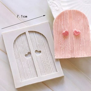 Könnte beinhalten: Eine weiße Silikonform zum Backen oder Basteln. Die Form ist wie eine Doppeltür mit einem gewölbten Oberteil und zwei herzförmigen Ausschnitten geformt. Die Form ist 7,1 Zoll breit.