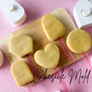 Puede incluir: Una tabla de madera muestra seis pasteles amarillos en palitos rosas. Los pasteles tienen varias formas: hexágono, corazón, círculo y rectángulos. El texto "Cakesicle Mold" está escrito en cursiva en la parte inferior.
