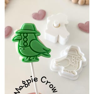May include: A green bird-shaped cookie on a stick, with a matching cookie cutter. The bird wears a hat and has three decorative circles. The words "Magpie Crow" are written in cursive. Two heart-shaped decorations are in the background.