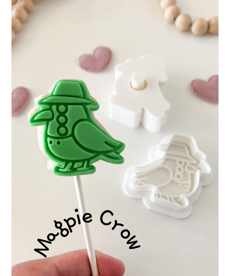 May include: A green bird-shaped cookie on a stick, with a matching cookie cutter. The bird wears a hat and has three decorative circles. The words "Magpie Crow" are written in cursive. Two heart-shaped decorations are in the background.
