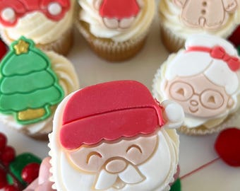 May include: Six decorated cupcakes with various Christmas themes. The cupcakes are topped with fondant figures of a Christmas tree, a gingerbread person, a girl in a red dress, a red truck, a woman with glasses, and Santa Claus.