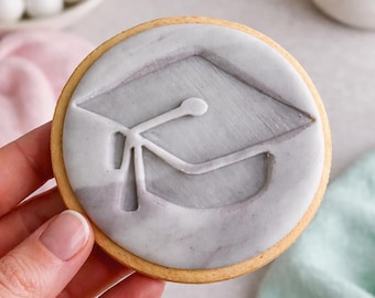 May include: A round sugar cookie featuring a smooth, light gray marbled icing layer. The surface is embossed with a clear, recessed outline of a graduation cap, complete with a tassel detail, centered on the cookie's top surface.