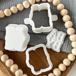 May include: A set of four white plastic cookie cutters shaped like a banner, a rectangle, and the letters "B" and "A".
