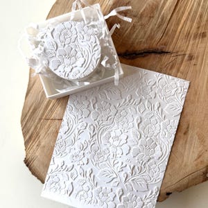 Puede incluir: Una tarjeta rectangular blanca y un disco circular, ambos con un patrón floral en relieve. Los artículos se presentan con papel triturado blanco dentro de una pequeña caja cuadrada, mostrando un diseño botánico consistente.