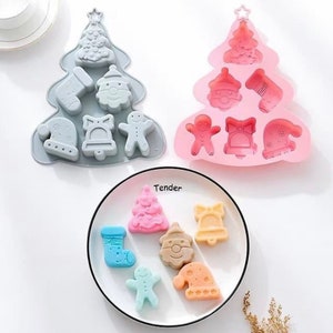 Op de afbeelding: Twee kerstboomvormige siliconen mallen, een grijze en een roze, met verschillende kerstthema-ontwerpen, waaronder een sok, een sneeuwpop, een bel, een peperkoekman en een kerstboom. Een bord met zes kerstthema-snoepjes gemaakt in de mallen.