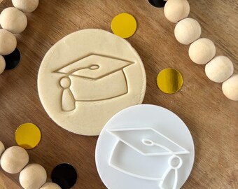 May include: A white plastic graduation cap cookie cutter on a wooden surface with a beige cookie dough imprint of the same design.