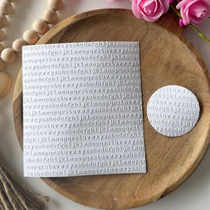 May include: A square and a round, light gray embossed alphabet design on a wooden tray. The square has rows of letters, and the round has a partial alphabet. Pink roses and wooden beads are in the background.