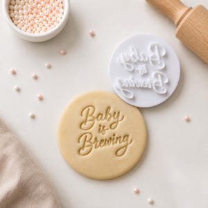 May include: A round cookie dough disc embossed with the text Baby is Brewing, accompanied by a white circular plastic stamp featuring the same raised lettering, a small bowl of light pink sprinkles, and a wooden rolling pin handle.