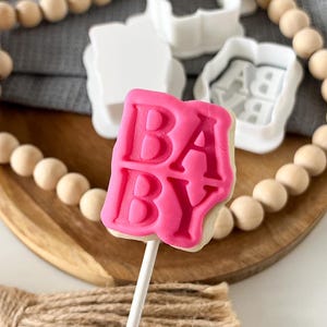 May include: Pink "BABY" cookie cutter on a stick with a wooden background and white cookie cutters in the background.