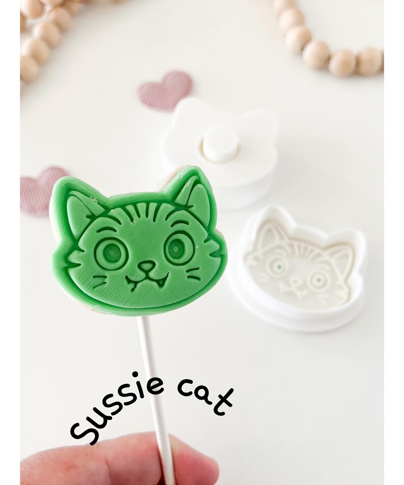 May include: A green cat-shaped lollipop on a white stick, with the text "Sussie cat". A white cat-shaped cookie cutter and a white stamp are also visible. Pink heart-shaped decorations and wooden beads are in the background.