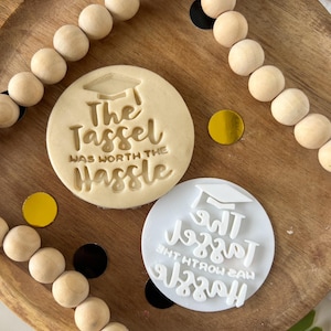 Puede incluir: Dos cortadores de galletas blancos con el texto "The Tassel Was Worth The Hassle" en negro. Los cortadores de galletas están sobre una superficie de madera con una cuerda de cuentas de madera.