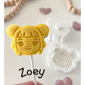May include: A yellow, cartoon-style cookie or candy on a stick, shaped like a girl's face, with the name "Zoey" written below. A white plastic mold and a pink heart-shaped decoration are also visible.
