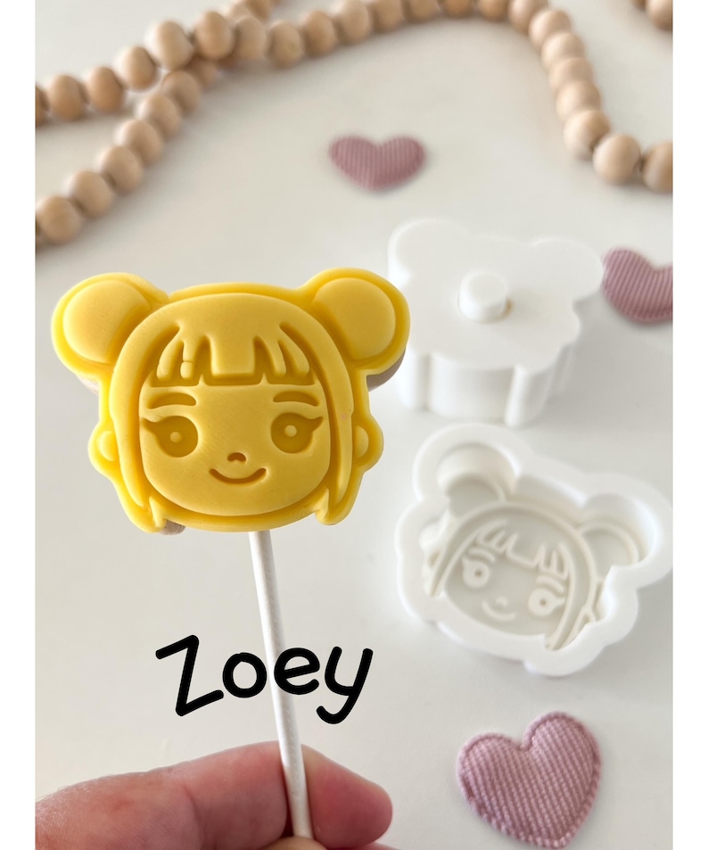 May include: A yellow, cartoon-style cookie or candy on a stick, shaped like a girl's face, with the name "Zoey" written below. A white plastic mold and a pink heart-shaped decoration are also visible.