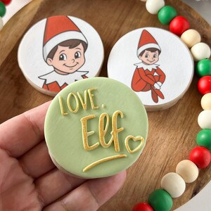 May include: Three round cookies with elf illustrations and a green cookie with the words "LOVE. Elf" in gold lettering. The cookies are on a wooden tray with a beaded garland in red, green, white, and beige.