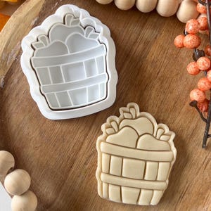 May include: A white plastic cookie cutter and a baked cookie in the shape of a basket filled with apples. The cookie cutter is on a wooden surface with decorative wooden beads and orange berries.