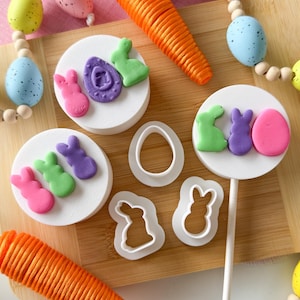 May include: An Easter-themed arrangement featuring colorful bunny and egg-shaped decorations on white circular platforms. A large orange carrot and beaded egg garlands are also present. White plastic bunny and egg-shaped cookie cutters are also visible.