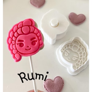 May include: A pink, cartoon-style character lollipop on a stick, with the name "Rumi" written in black script. Also shown are a white cookie cutter, a matching stamp, and three small, pink heart-shaped fabric decorations.