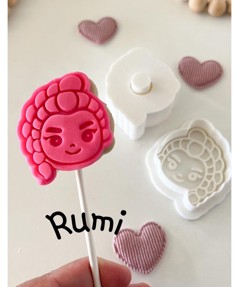May include: A pink, cartoon-style character lollipop on a stick, with the name "Rumi" written in black script. Also shown are a white cookie cutter, a matching stamp, and three small, pink heart-shaped fabric decorations.