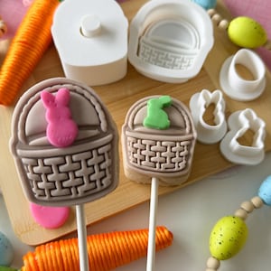 May include: An assortment of Easter-themed baking supplies and treats. Two basket-shaped lollipops with bunny accents sit on a wooden board, alongside carrot and egg decorations. White plastic molds and cutters are also visible, suggesting a DIY baking project.