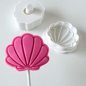 May include: A vibrant pink seashell lollipop on a white stick, alongside two white seashell-shaped cookie cutters. One cutter is a complete shell shape, and the other is a flower shape with a small cylinder in the centre.
