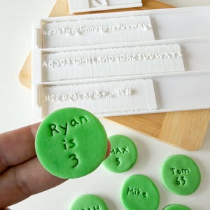 Peut inclure: Un ensemble de tampons de lettres et de chiffres en plastique blanc avec une planche à découper en bois. L'ensemble comprend des tampons de lettres et de chiffres individuels, et un cercle de fondant vert avec le nom "Ryan", le mot "is", et le chiffre "3" estampillé dessus.