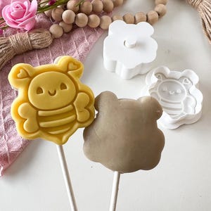 以下が含まれることがあります： 白い棒に刺さった黄色い蜂の形をしたケーキポップと、薄茶色のケーキポップ。白い蜂の形をしたクッキーカッターも2つ見えます。背景にはピンクの花、木のビーズ、ピンクの布が含まれています。
