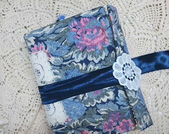 May include: A blue and pink floral fabric covered journal with a navy blue ribbon and a white lace trim. The journal has a white lace flower embellishment.