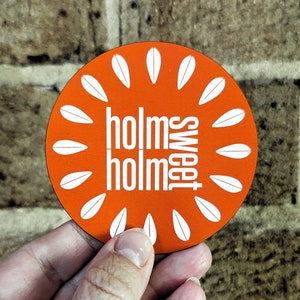 May include: A round orange coaster with the words "holms holm sweet" in white. The coaster has a white petal design around the edge. The coaster is held in a hand against a brick wall.