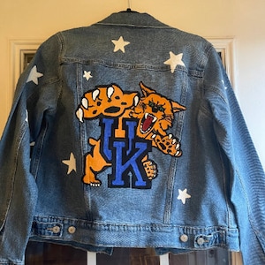 May include: A blue denim jacket with white stars and a large orange and blue patch of a wildcat with the letters "UK" on it.