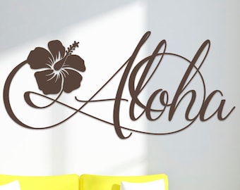 Pode incluir: Um adesivo de parede de vinil marrom com a palavra "Aloha" em uma fonte cursiva com uma flor de hibisco marrom à esquerda da palavra. O adesivo está em uma parede branca acima de um sofá amarelo com almofadas brancas.