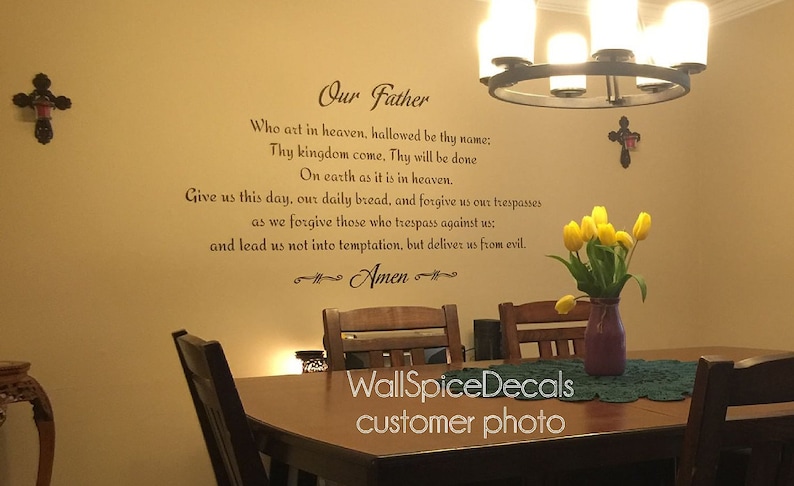 May include: A dining room setting with a wall decal displaying the Lord's Prayer. The room includes a wooden table, chairs, and a vase of yellow tulips. Decorative crosses are on the walls, and a chandelier hangs from the ceiling.