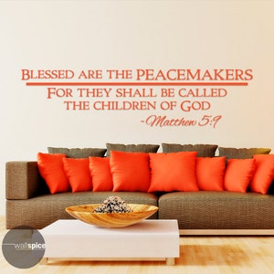 May include: A wall decal with the Bible verse "Blessed are the peacemakers, for they shall be called the children of God - Matthew 5:9" in orange text on a white wall. The decal is displayed above a brown couch with orange throw pillows.